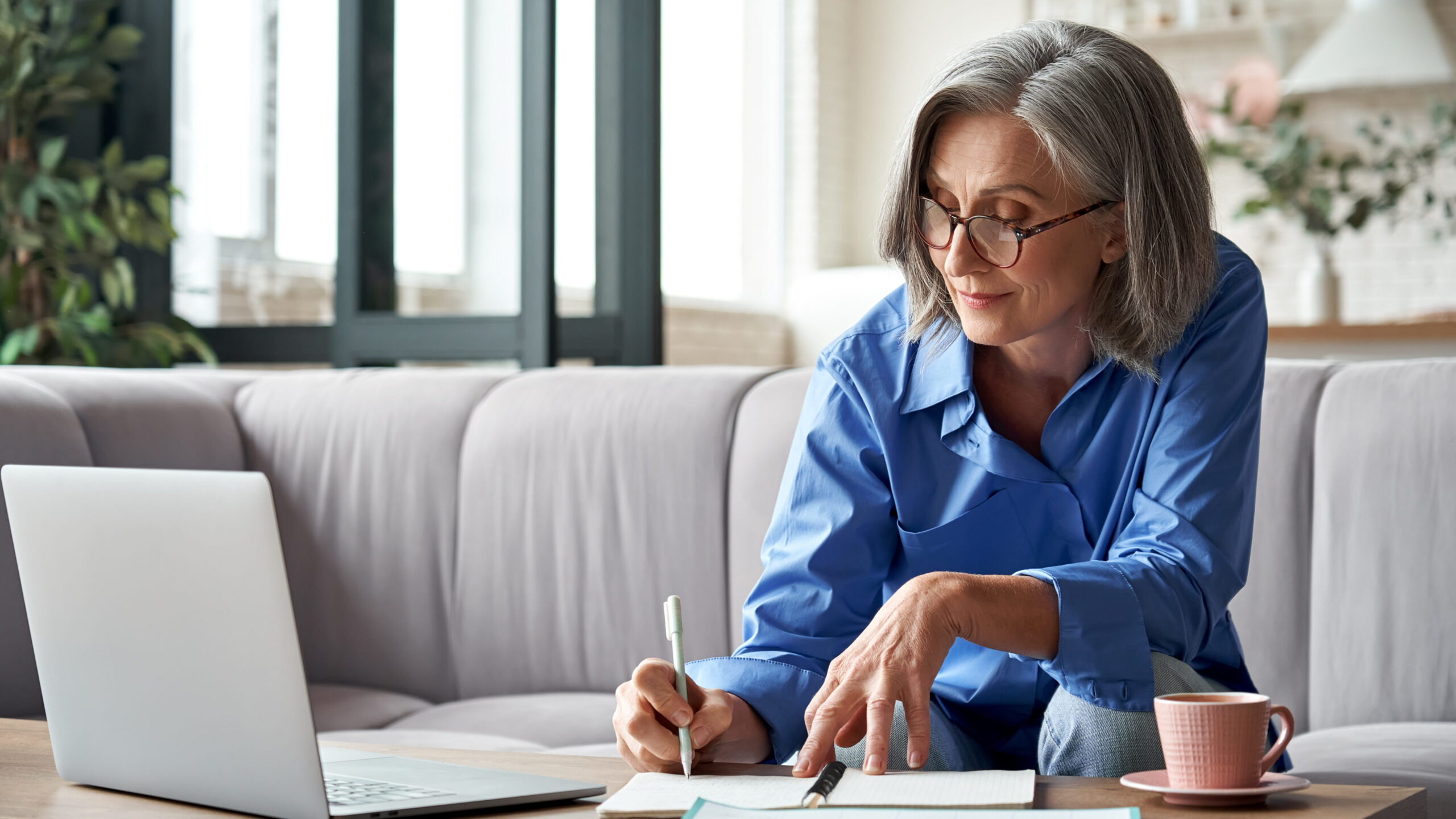 Stylish mature older woman working from home on laptop taking notes.