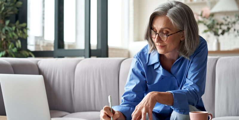 Stylish mature older woman working from home on laptop taking notes.