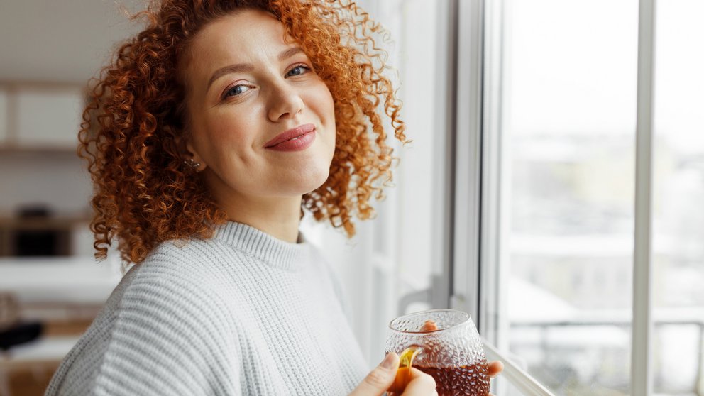 Closeup image of cute attractive millennial redhead woman with funny curls enjoying tea standing next to big panoramic window with city landscape, looking at camera with happy satisfied smile