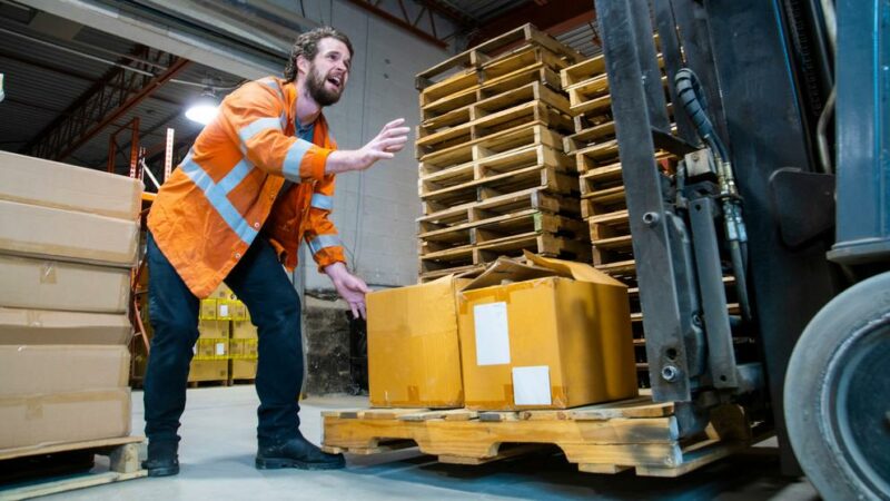 An industrial warehouse workplace safety topic. A forklift driver cause serious injury as he lowers his forks onto a coworker's foot.