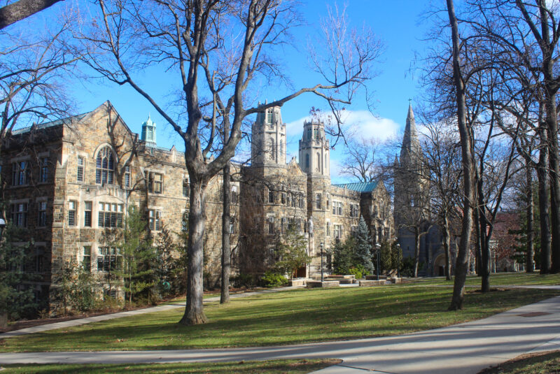A multi-story stone building with two prominent towers and a church steeple in the background, set against a bright blue sky with some clouds.